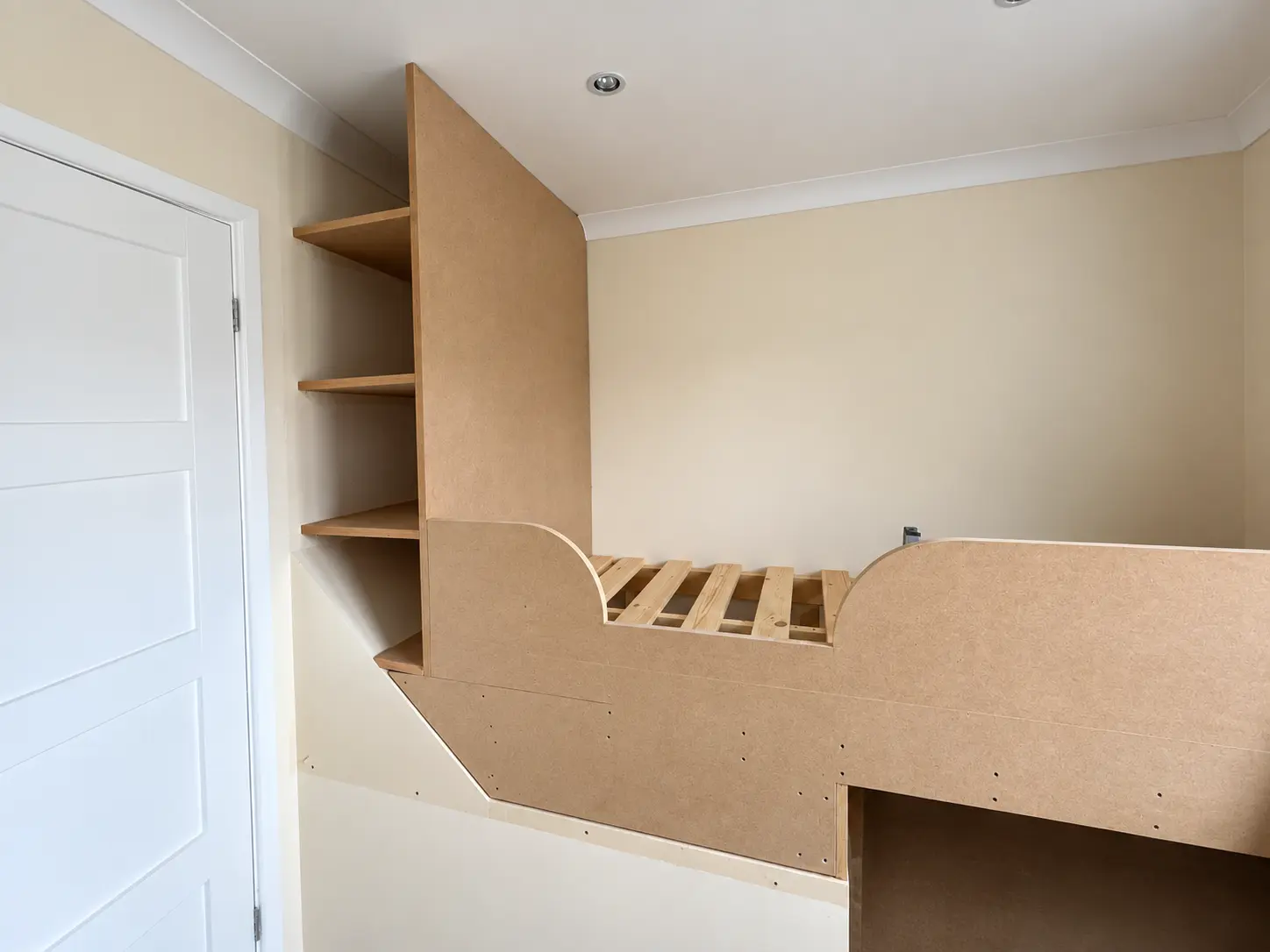 Unfinished bespoke MDF raised bed with integrated shelving beside the bedroom door